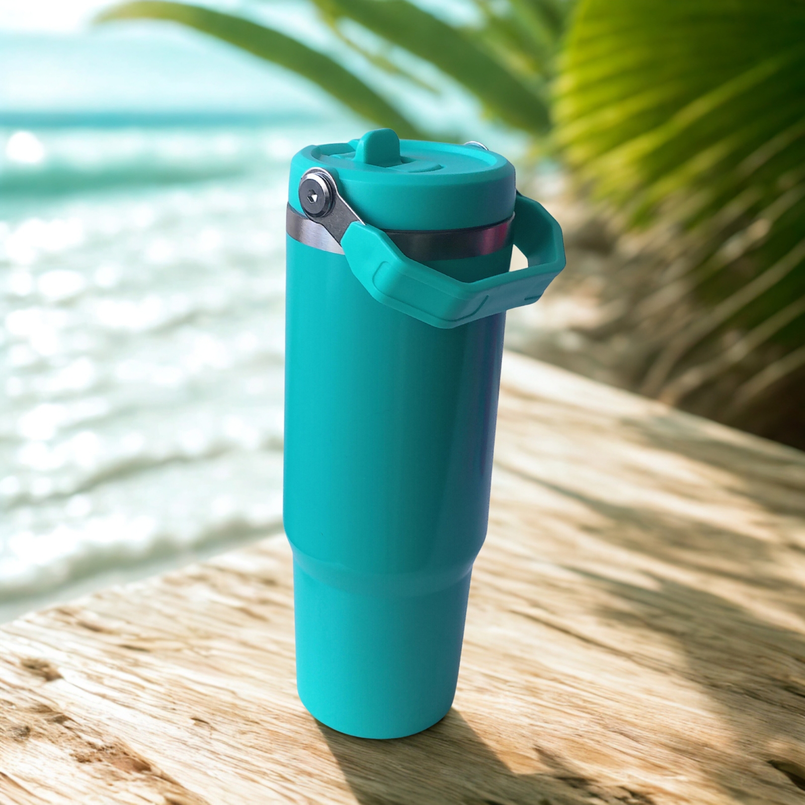 Angled side view of the Tropical Turquoise tumbler with lid closed and handle down, placed on a wooden deck beside the ocean